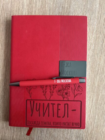 Red hardcover notebook with floral illustration and bold sign-off on the bottom, a red pen resting across its cover, placed on a wood surface.